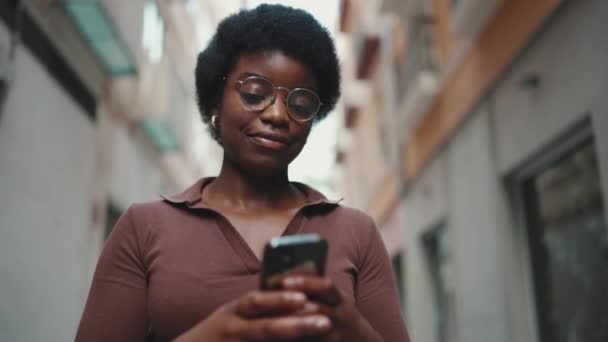 スマートフォンを使って通りを歩いている若いアフリカ人の少女 可愛いです女性チェック彼女の携帯電話屋外 — ストック動画