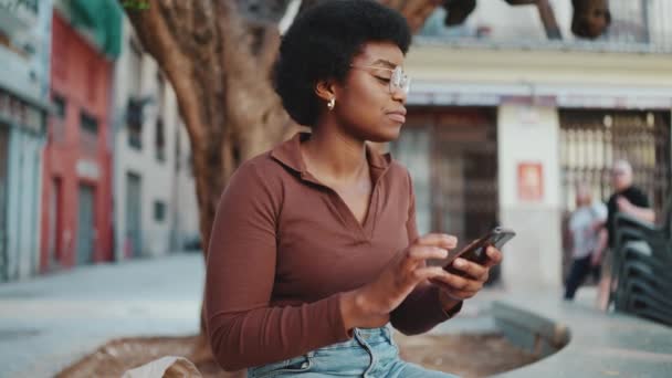 魅力的なアフリカの女性が屋外で電話で友人とチャット かなり暗いです肌の女の子チェック彼女のスマートフォン上の通り — ストック動画