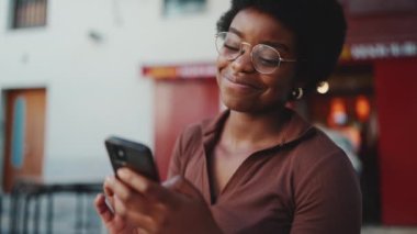 Gözlüklü, akıllı telefondan mesaj okuyan ve gülümseyen güzel bir Afrikalı kız. Dışarıda cep telefonu kullanan genç bir kadın.