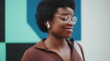 Gözlüklü Afrikalı kadın, şehirde yürürken kablosuz kulaklıkla müzik dinlerken ve dans ederken görülüyor. Kaygısız kız