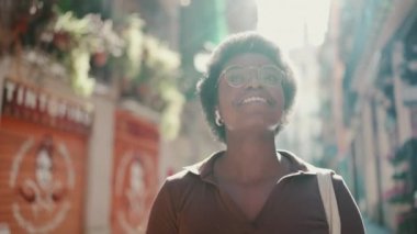 Güzel, kıvırcık saçlı, gözlüklü, kablosuz kulaklıkla müzik dinleyen Afro-Amerikan bir kadın. Afrikalı kız mutlu bir şekilde sokakta yürüyor.