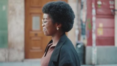 Ceket giymiş, sokakta yürürken gülümseyen Afrikalı bir kadının yan görüntüsü. Afro-Amerikalı kız dışarıda güzel bir gün geçiriyor.
