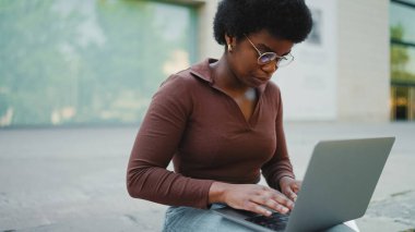 Üniversite kampüsünde dizüstü bilgisayar kullanan Afrikalı öğrenci. Genç kıvırcık kadın dışarıda dizüstü bilgisayarla çalışıyor.
