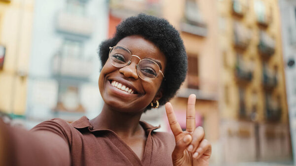 Positive African American dark haired woman taking selfie on the street. Pretty girl showing peace gesture on camera