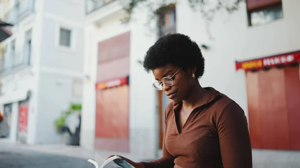 African American woman in glasses sitting on the street and reading a textbook. Female dark-skinned student studying outdoors