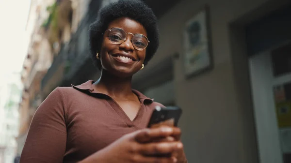 Cheerful African woman in glasses holding smartphone on the street. Girl looking happy walking on street and using her mobile phone