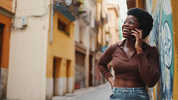 Pretty dark-skinned female in glasses talking over mobile phone. Cheerful African woman wearing casual having a conversation on phone outdoors