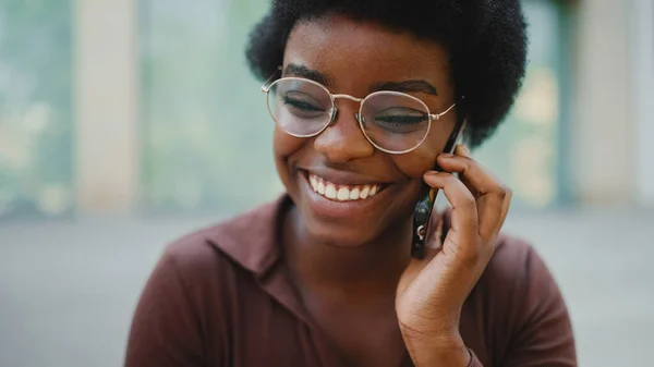 Pretty dark-skinned female in glasses having conversation with friend on mobile phone. African woman looking happy talking over mobile phone outdoors