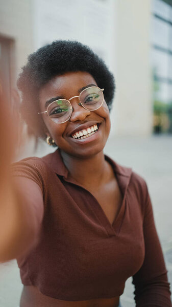 Cheerful African American curly haired girl looking cute taking selfie outdoors. Dark-skinned woman in glasses looking happy sincerely smiling at camera