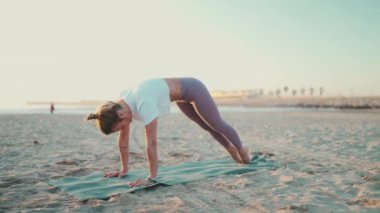 Havalı bir kadının deniz kenarındaki spor minderinde yoga yaptığı güzel bir kare. Spor kıyafetleri giymiş, kumsalda köpek pozu veren sarışın yogi kızı.