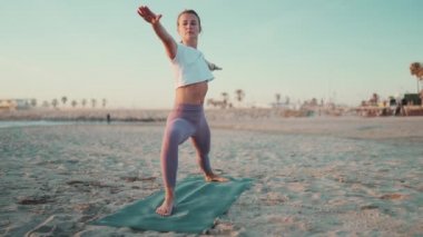 Güzel bir kızın plajdaki spor minderinde yoga yaptığı uzun bir çekim. Spor giyimli çekici bir kadın yoga kursu sırasında dışarıda esneme egzersizi yapıyor.
