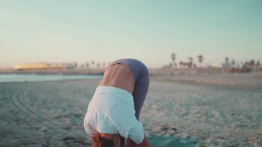 Spor elbiseli genç bir kadın plajda yoga egzersizi yapıyor. Güzel kız dışarıda esneme egzersizleri yapıyor. Yoga kızı.