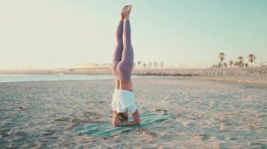 Yoga pozu veren güçlü bir Yogi kızı inanılmaz bir şekilde kafasının üstünde duruyor. Çekici kadın sahilde sabah yogası yapıyor.