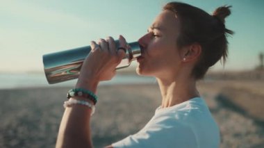 Kumsalda yoga yaparken su içen çekici sarışın kadın. Spor dövmeli kız deniz kenarında dinleniyor. Su dengesi