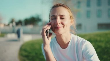 Sarışın, mutlu görünen, akıllı telefondan konuşan, şehir parkında egzersiz yapmayı bekleyen bir kadın. Uzak iletişim