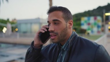 Gündelik giyinmiş genç iş adamının akşam vakti akıllı telefondan konuşurken çekilmiş görüntüsü. Dışarıda cep telefonuyla konuşan çekici bir adam.