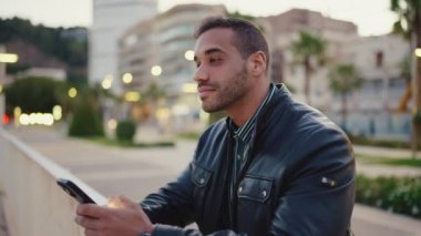 Deri ceketli genç esmer adam dışarıda akıllı telefonunu kontrol ediyor. Sokakta arkadaşlarını beklerken akıllı telefon kullanan çekici bir adam.