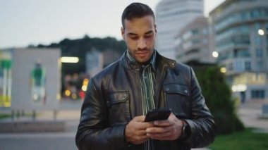 Deri ceketli genç adam dışarıda akıllı telefonunu kontrol ediyor. Cep telefonlu çekici bir adam akşam kentinde yürüyor.