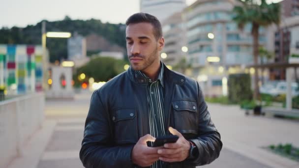 年轻的黑发男人在外面等女朋友的时候 用智能手机看起来很沉思 迷人的小伙子一个人走在傍晚的城市里 — 图库视频影像