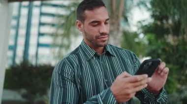 Tişört giymiş, kendine güvenen iş adamını internette çalışırken akıllı telefonu kullanarak kapat. Güzel adam dışarıda akıllı telefonunu kontrol ediyor.