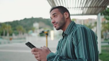 Sokakta oturan ve akıllı telefonunu kontrol eden gömlekli çekici bir adamın yan görüntüsü. Esmer adam dışarıda cep telefonu kullanıyor.