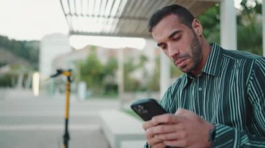 Çizgili tişörtlü genç adamın sokakta oturup akıllı telefonunu kontrol edişinin görüntüsü. Çekici bir adam cep telefonu kullanıyor ve dışarıda gülümsüyor.