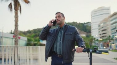 Genç bir adamın akıllı telefondaki arkadaşıyla sokakta e-scooterlı bir şekilde konuşurken görüntüsü. Çekici adam dışarıda elinde telefon ve elektrikli scooterla mutlu görünüyor.