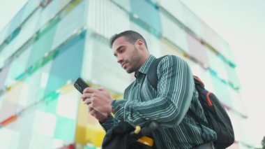 Sırt çantalı genç adam sokakta e-scooter kiralamak için akıllı telefon kullanıyor. Çekici iş adamı scooter üzerinde çalışacak.