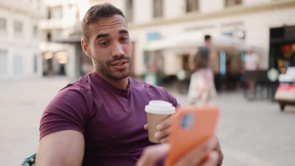 漂亮的黑发小伙子在街上咖啡店休息的时候和朋友有一个视频通话 小伙子在户外用智能手机讲话时 随便说说 把咖啡拿走了 — 图库视频影像