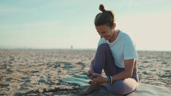 Piękna Kobieta Siedząca Macie Jogi Sprawdzająca Smartfona Plaży Młoda Kobieta Obrazy Stockowe bez tantiem