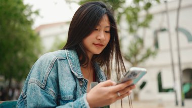 Uzun saçlı, kot ceket giyen, açık havada akıllı telefonunu kontrol eden çekici bir Asyalı kızın portresi. Cep telefonlu kız öğrenci bankta dinleniyor.