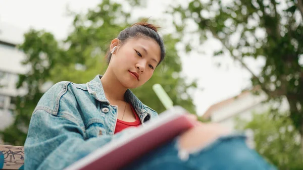 美丽的亚洲女孩 戴着无线耳机 坐在大学校园里 在书上记笔记 女生在户外做作业 — 图库照片