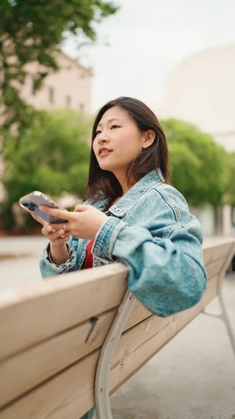 Ganske Langhåret Asiatisk Kvinne Med Dongerijakke Sittende Benken Med Smarttelefon – stockfoto