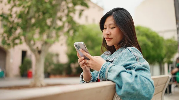 美丽的长发亚洲女孩穿着斜纹棉布夹克在户外查看她的社交媒体 女学生拿着智能手机躺在长椅上 — 图库照片