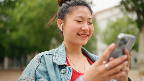 スマートフォンをチェックし 屋外で微笑むワイヤレスイヤフォンを身に着けている喜びを探しているかなりアジアの女性 近代技術の概念 ロイヤリティフリーのストック写真