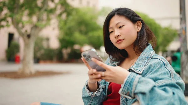 Langhåret Asiatisk Kvinne Med Dongerijakke Sittende Benken Utendørs Sjekker Smarttelefonen – stockfoto