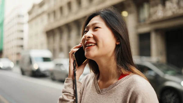 Pretty Asian woman standing on the street talking on smartphone with her friend telling what happened today. Asian girl looking happy using mobile outdoors