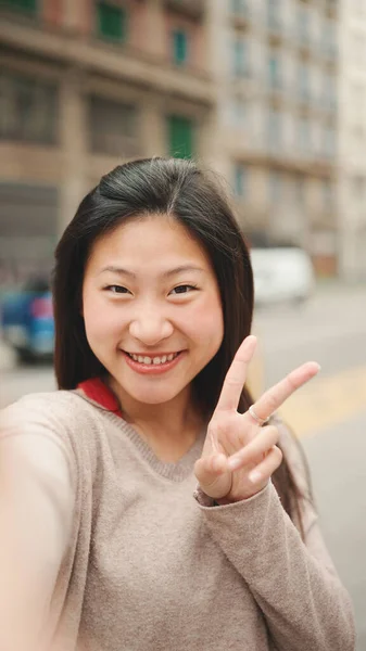 Pretty Asian girl taking self portrait showing peace sign outdoors. Long haired smiling woman posing on the street