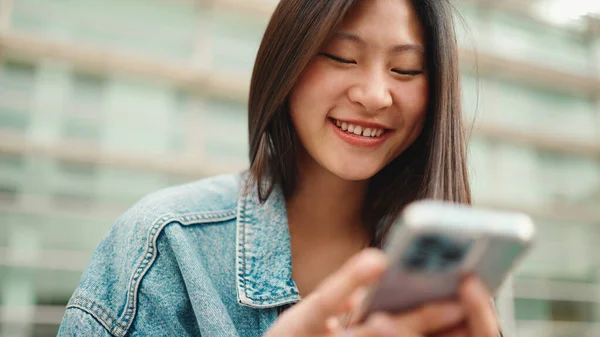 Fröhliches Langhaariges Asiatisches Mädchen Das Draußen Mit Dem Smartphone Lacht Stockbild