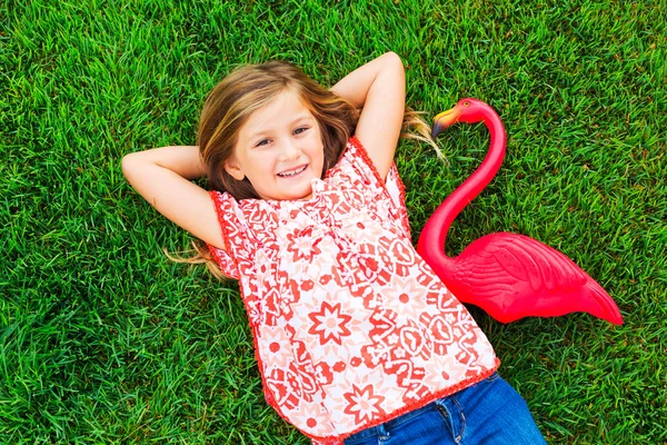 Smiling little girl lying on green grass with pink flamingo — Stock Photo, Image