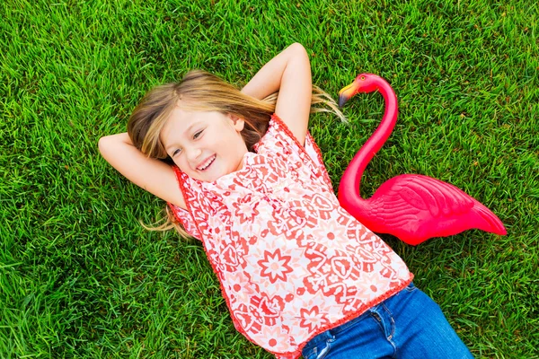 Улыбающаяся маленькая девочка лежит на зеленой траве с розовым фламинго — стоковое фото