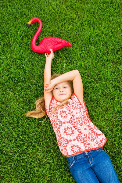 Sorridente bambina sdraiato sull'erba verde con fenicottero rosauśmiechnięta dziewczynka leżący na zielonej trawie z różowy Flaming — Zdjęcie stockowe