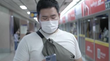 Maske takan Asyalı mutlu bir adam Covid19 Omicron 'u korumak için cep telefonu kullanıyor metro trenini bekliyor.