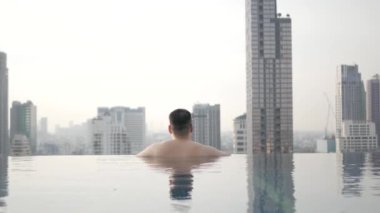 Asyalı adam gün batımında şehrin arka planında sonsuz yüzme havuzunda dinlenirken