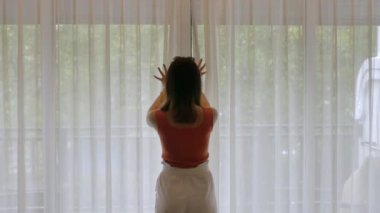 Asyalı kadın yatak odasındaki beyaz perdeyi açtı. Dışarıdaki doğal ağaç ve balkon manzaralı.