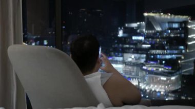 View to asian man use hand grap a glass of water to drink while relaxing on sofa in hotel living room in night time