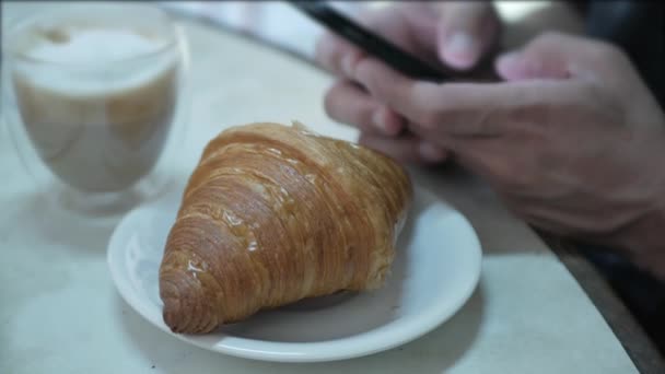 一名男子使用手机社交媒体 从盘子里抓起新鲜的法国羊角面包 有选择地把重点放在早餐或下午茶小吃上 — 图库视频影像