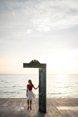 Kadın balerin, güneş doğarken deniz kenarında sanat objesinin yanında poz veriyor.
