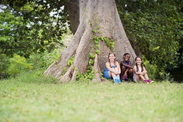 Tres amigas felices sentadas cerca de un gran árbol — Foto de Stock