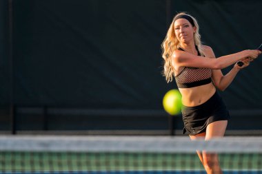 Sarışın bir model tenis kortunda öğleden sonra güneşinde tenis oynuyor.